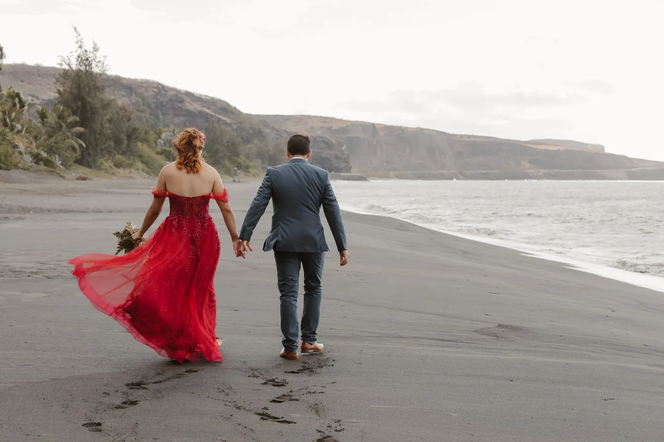 Les mariés marchent main dans la main sur une plage de sable noir, la robe rouge au vent