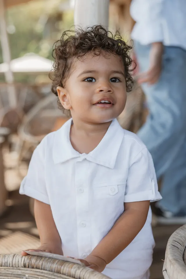 Un petit garçon aux cheveux bouclés, en chemise blanche, sourit appuyé sur un fauteuil en rotin