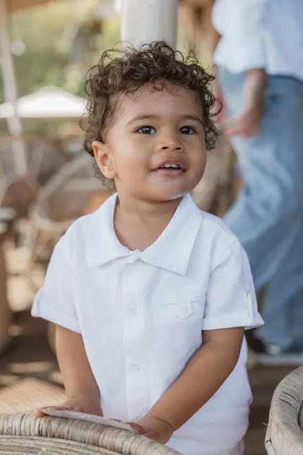 Un petit garçon aux cheveux bouclés, en chemise blanche, sourit appuyé sur un fauteuil en rotin