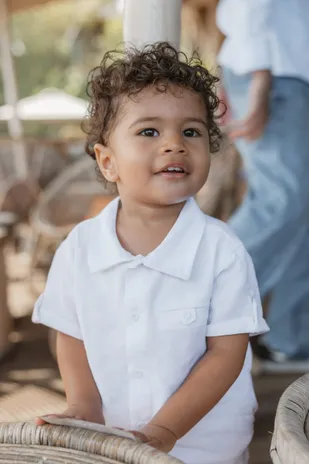 Un petit garçon aux cheveux bouclés, en chemise blanche, sourit appuyé sur un fauteuil en rotin