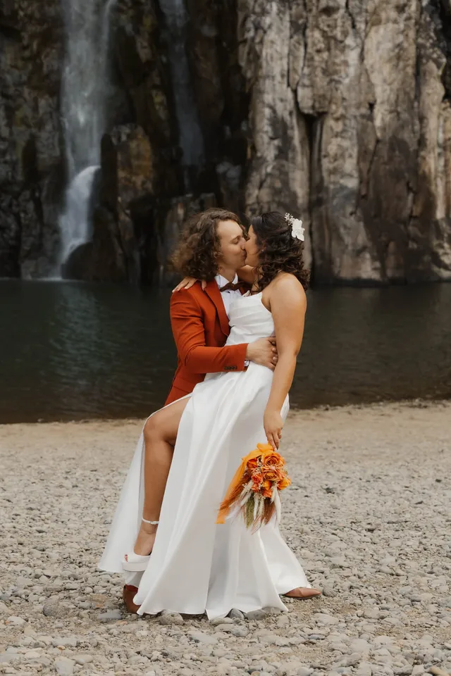 Le marié en veste rouille soulève la mariée pour un baiser, devant la cascade