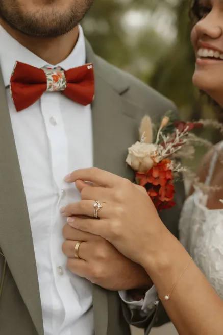 La main de la mariée, bague au doigt, posée sur la veste du marié au nœud papillon rouille
