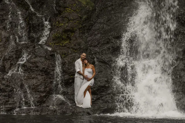 Un couple enlacé au pied d’une cascade, les mains posées sur le ventre rond