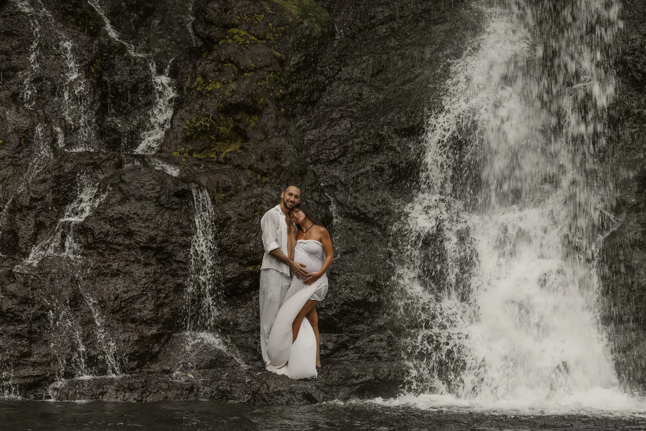 Un couple enlacé au pied d’une cascade, les mains posées sur le ventre rond