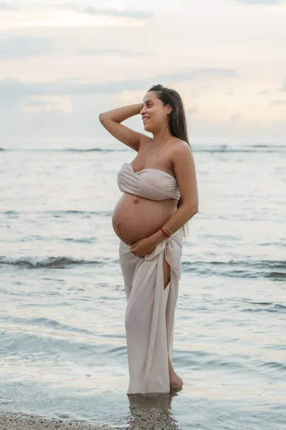 Future maman drapée de beige debout dans l’eau, main dans les cheveux, regard vers le large