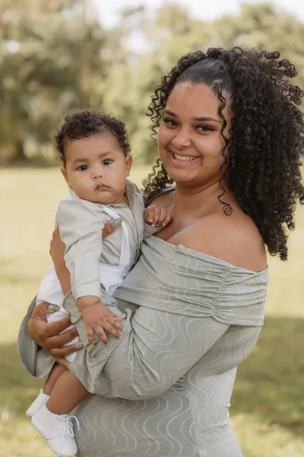 Jeune femme souriante portant l’enfant en tenue claire sur sa hanche, dans un jardin