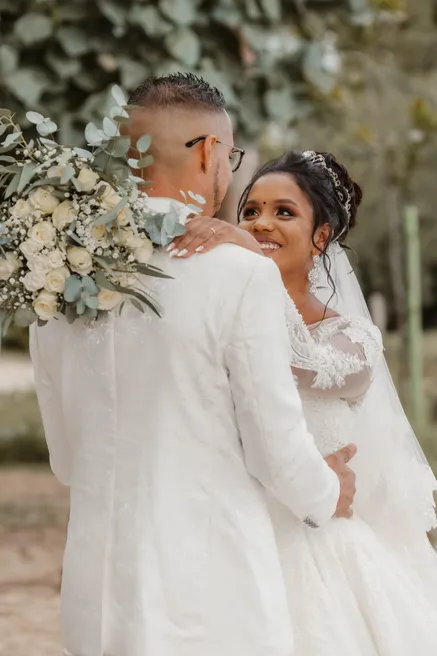 Le marié en costume blanc enlace la mariée voilée, bouquet blanc et eucalyptus sur son épaule