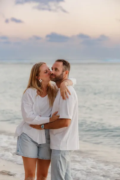 Un couple s’enlace au bord de l’eau, elle l’embrasse sur la joue sous un ciel rosé
