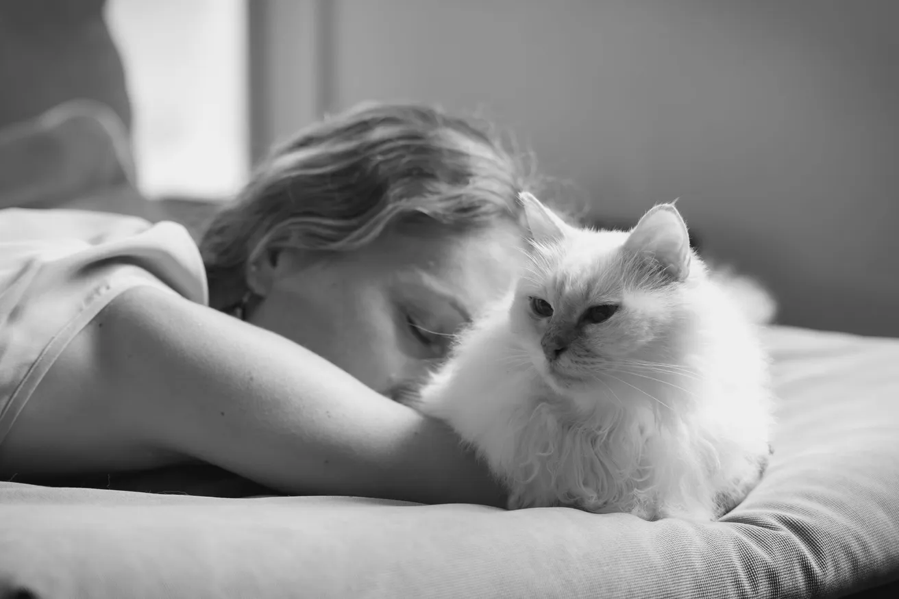 En noir et blanc, une femme allongée sur un lit ferme les yeux contre son chat à poils longs