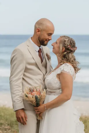 Les mariés front contre front au bord de l’océan, la mariée tenant un bouquet de fleurs séchées