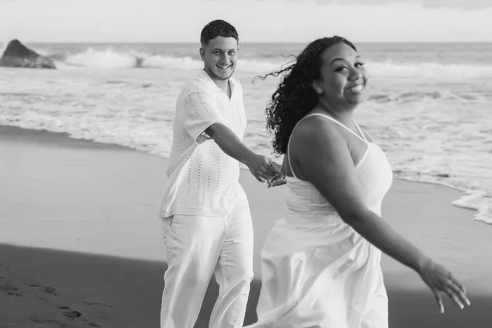 En noir et blanc, un couple marche en riant sur une plage de sable noir