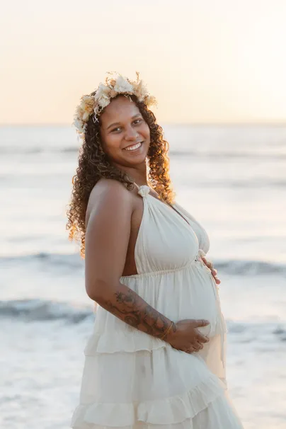 Future maman souriante devant la mer, couronne de fleurs séchées et robe crème