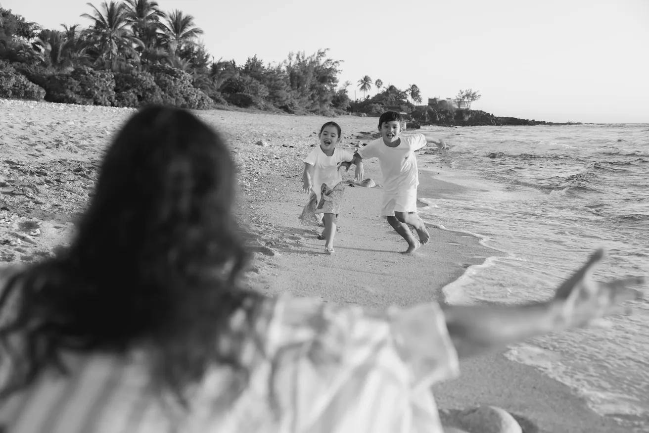 En noir et blanc, deux enfants courent sur le sable vers une femme de dos au premier plan