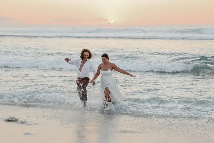 Les mariés courent dans l’écume au coucher du soleil, la mariée soulève sa robe