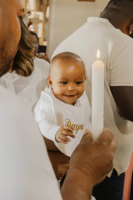 Un bébé en tenue blanche sourit à côté du cierge de baptême tenu par un proche