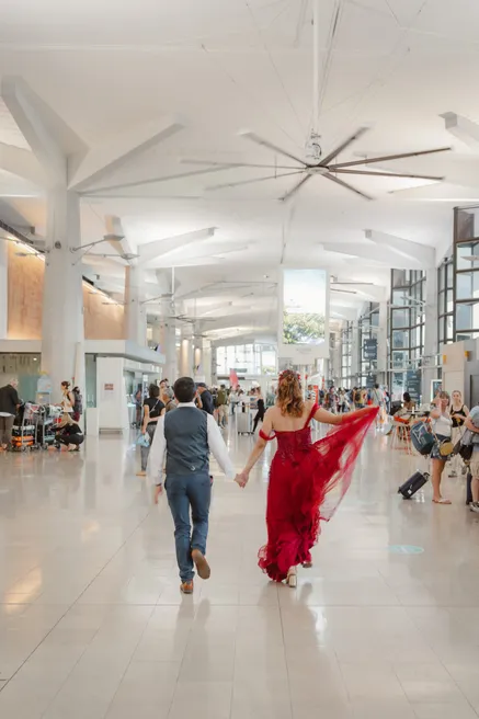 Les mariés marchent main dans la main dans un hall d’aéroport, la robe rouge au vent