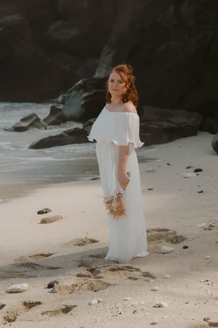 La mariée debout seule sur une plage de sable, robe longue et rochers sombres derrière elle
