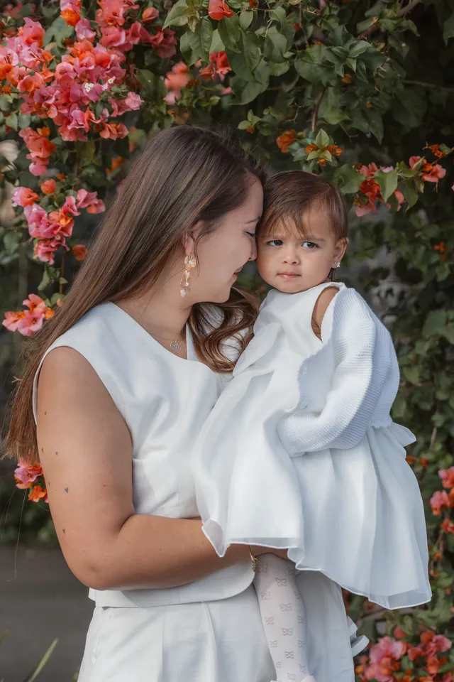 Une maman en blanc embrasse sa petite fille en robe de baptême, devant les bougainvilliers
