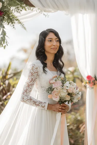 La mariée pose près d’un voilage blanc, voile long et bouquet de roses pâles