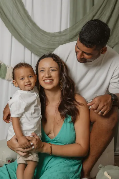 Un papa et une maman sourient, leur petit garçon en chemise blanche dans les bras, devant le décor sauge