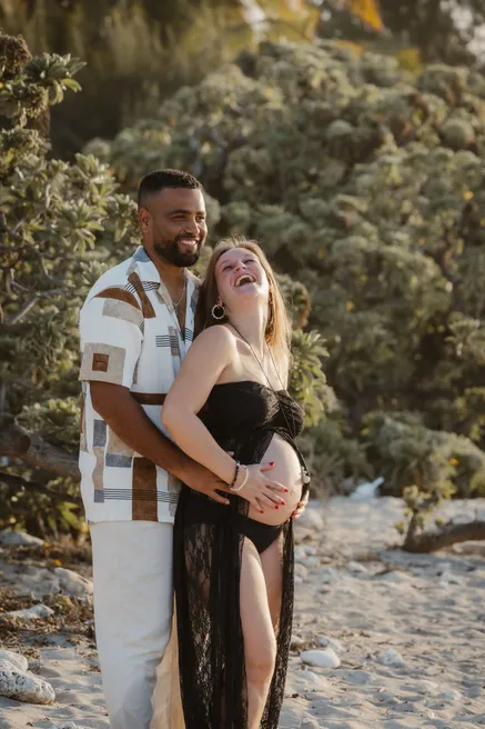 Un couple enlacé en riant sur la plage, la future maman en body noir et voile de dentelle