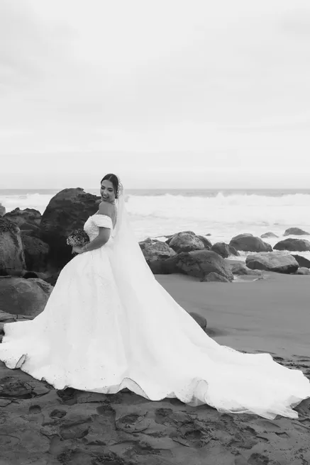 En noir et blanc, la mariée pose sur une plage de galets, traîne étalée sur le sable