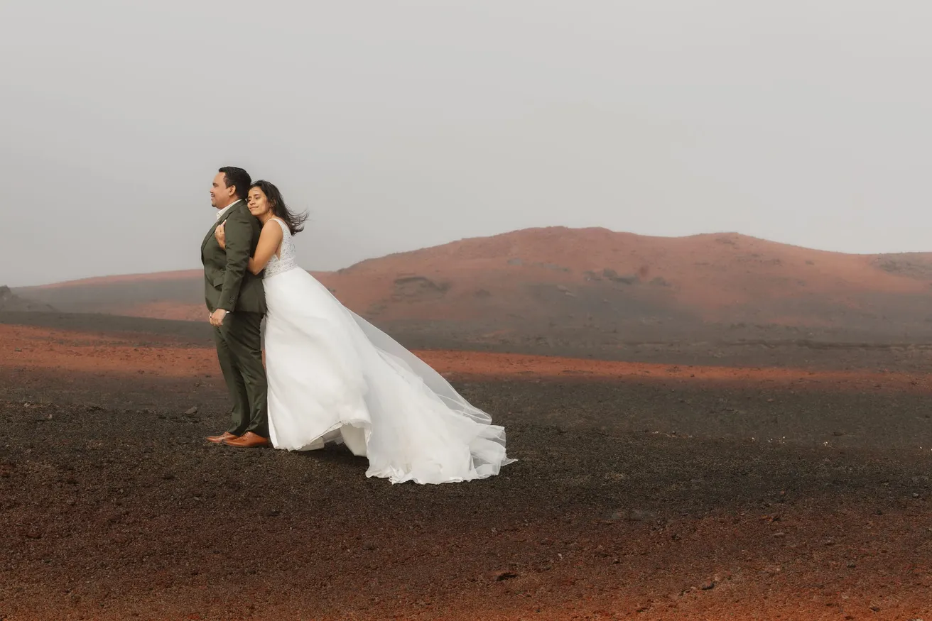 Blottie dans le dos du marié, la mariée pose dans le vent, sa robe soulevée, devant un cône volcanique rouge