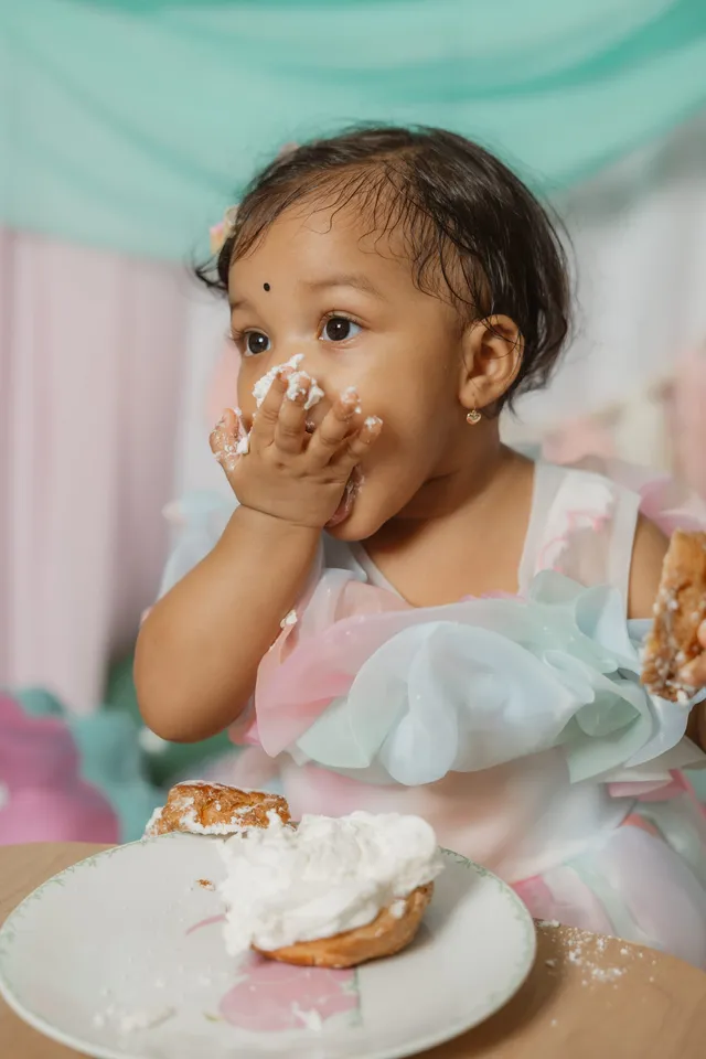 Une petite fille, les joues pleines de crème, porte les mains à sa bouche devant son assiette de gâteau