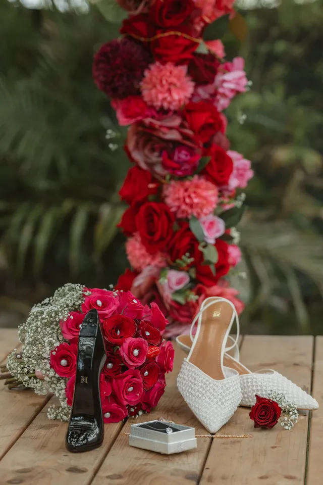 Escarpins, bouquet de roses rouges et écrin des alliances posés sur une table en bois, sous une arche de fleurs