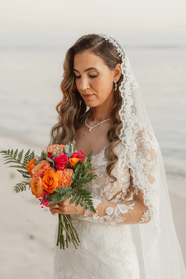 La mariée, voile de dentelle et bouquet orange, baisse les yeux devant la mer