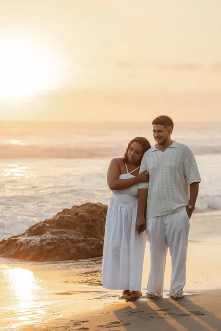 Un couple en blanc pose près d’un rocher sur le sable, dans la lumière du soleil couchant