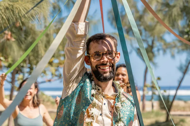 Le marié en veste turquoise brodée lève le bras sous les rubans colorés, collier de fleurs au cou