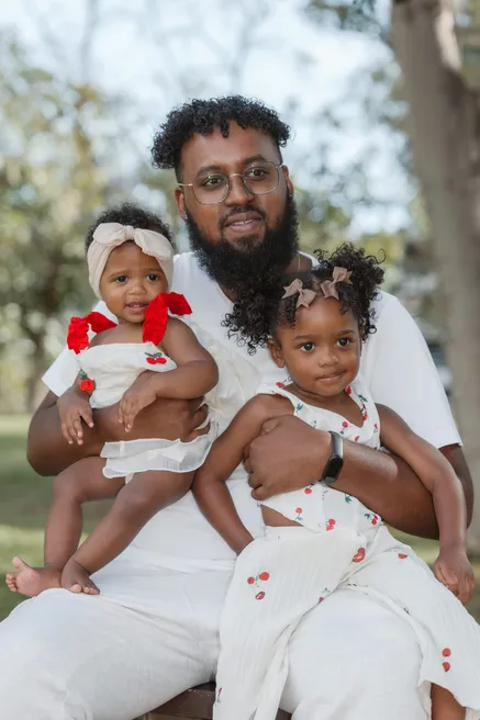 Un père assis sourit avec ses deux petites filles sur les genoux, en tenues à cerises assorties