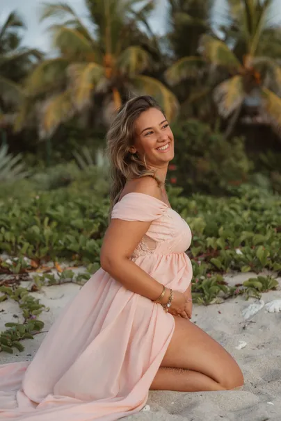 Future maman en robe rose poudré, agenouillée sur le sable, riant, palmiers derrière