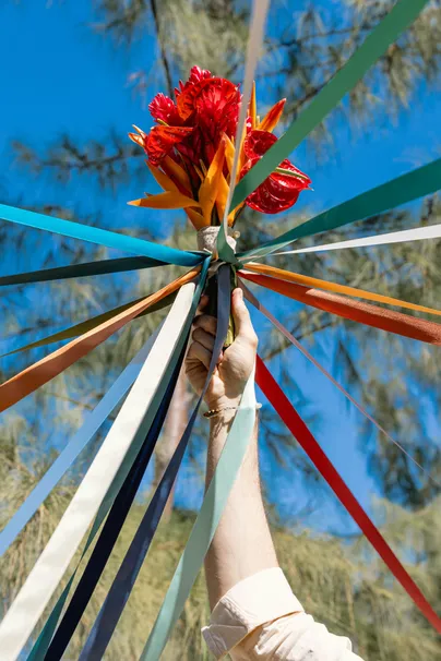 Rubans colorés noués à un mât sous un ciel bleu, une main tient des fleurs rouges au sommet