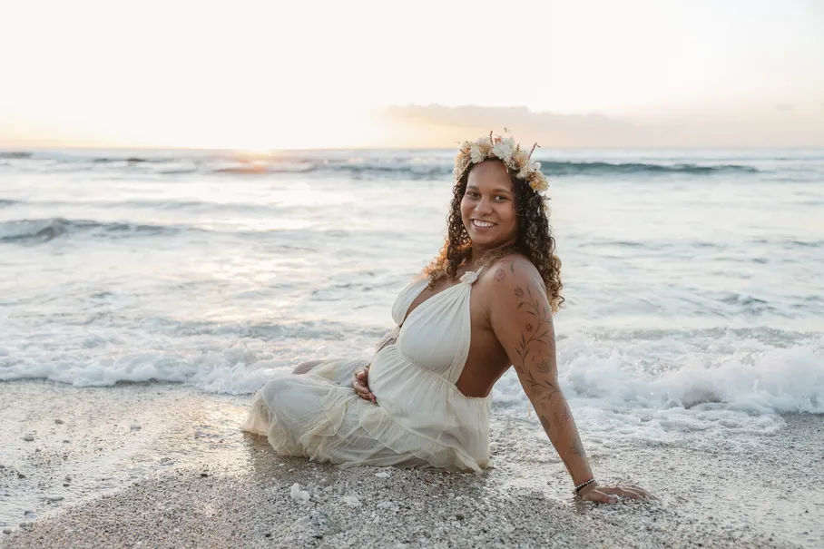 Future maman couronnée de fleurs assise sur le sable au bord de l’eau, souriante