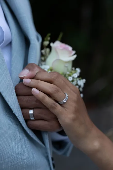 Mains des mariés jointes contre la veste bleu clair, alliances et boutonnière de rose
