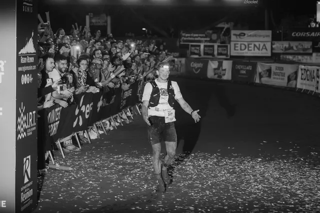 En noir et blanc, un coureur franchit la ligne d’arrivée de nuit, salué par la foule derrière les barrières