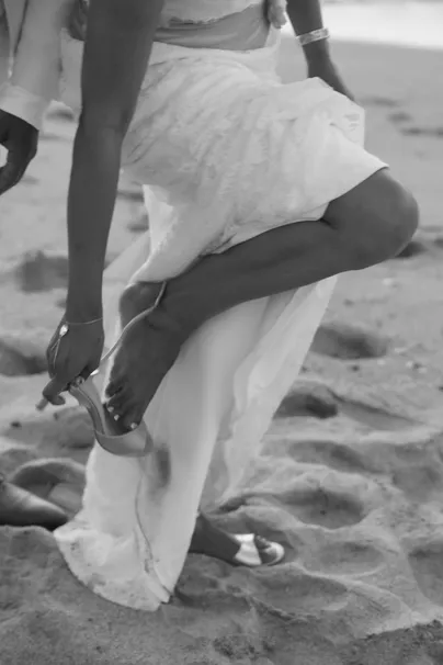 En noir et blanc, la mariée relève sa robe pour attacher sa sandale sur le sable