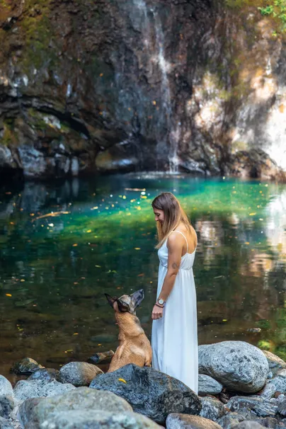 Une femme en robe blanche debout au bord d’un bassin d’eau verte, son chien à ses pieds, devant la cascade