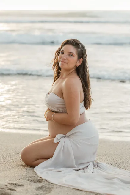 Future maman agenouillée sur le sable, jupe blanche étalée, sourire vers l’objectif
