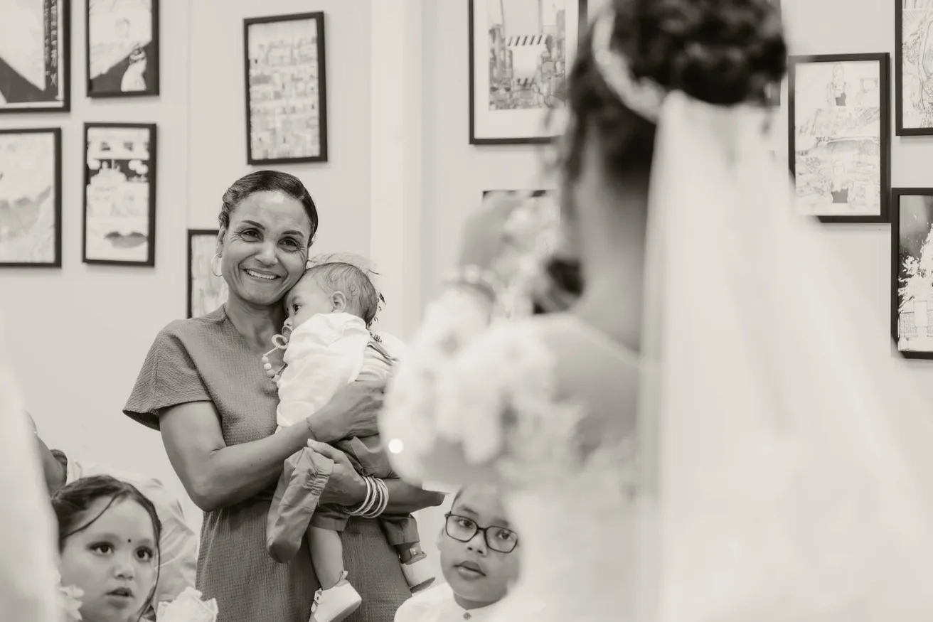 En noir et blanc, une femme tient un bébé parmi des enfants, près du voile de la mariée