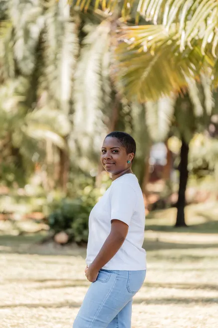 Une femme en tee-shirt blanc et jean, de trois quarts, sourit dans un jardin de palmiers