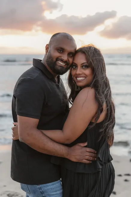 Un couple enlacé sourit face à l’objectif, devant la mer et les nuages du soir