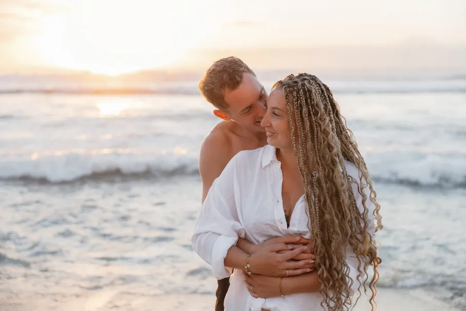 Un couple enlacé dans les vagues au coucher du soleil, il l’embrasse sur la joue