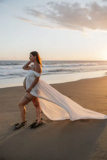 Future maman en robe blanche marchant sur le sable, longue traîne soulevée par le vent
