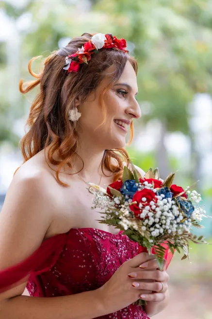 La mariée en robe bustier rouge et couronne de fleurs sourit, bouquet bleu, blanc et rouge à la main