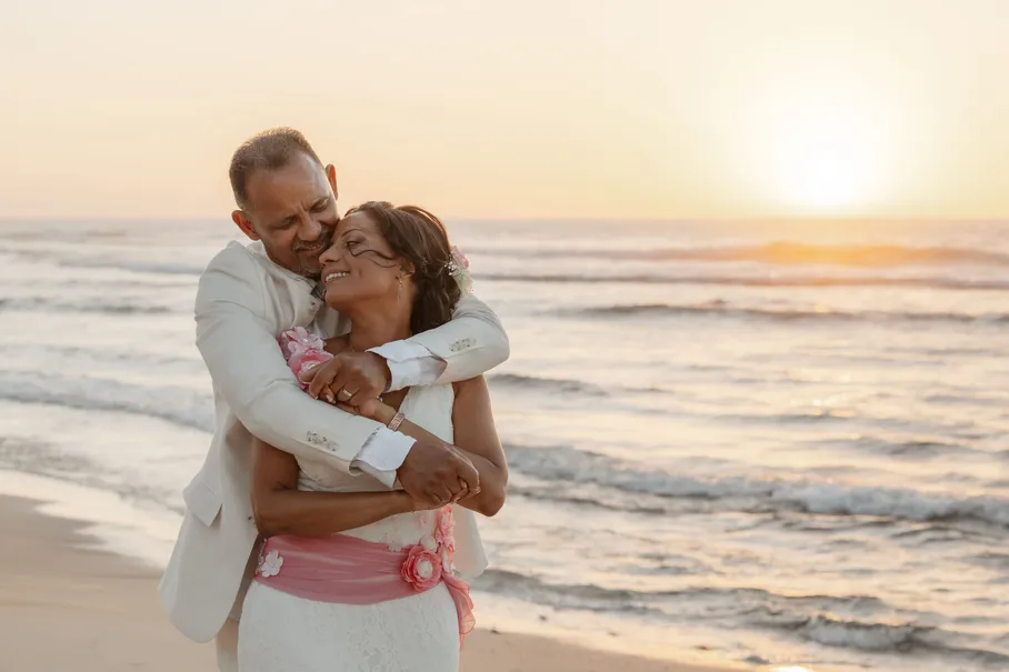 Le marié enlace la mariée par derrière sur la plage, le soleil couchant sur la mer