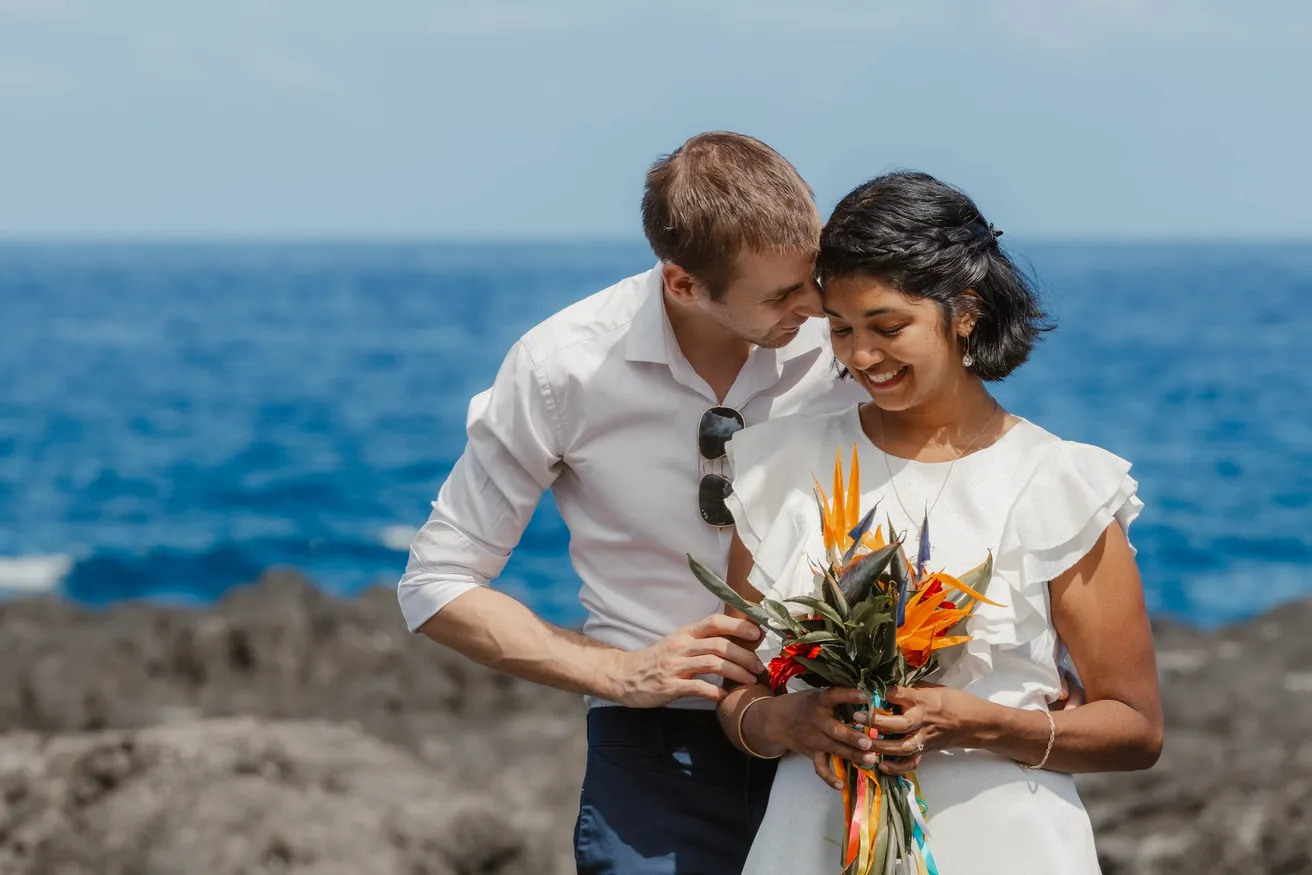 Un couple rit front contre front devant l’océan, elle tient un bouquet d’oiseaux de paradis