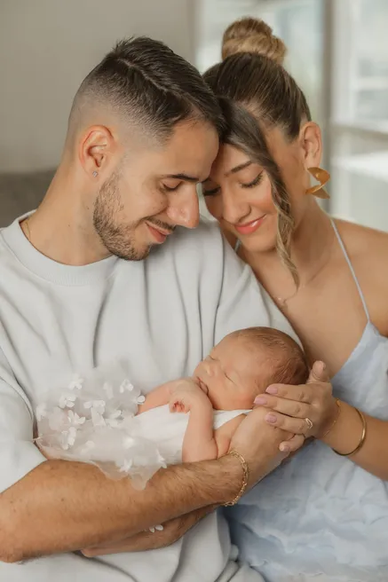 Un couple penché l’un vers l’autre tient son nouveau-né en tenue ornée de fleurs blanches